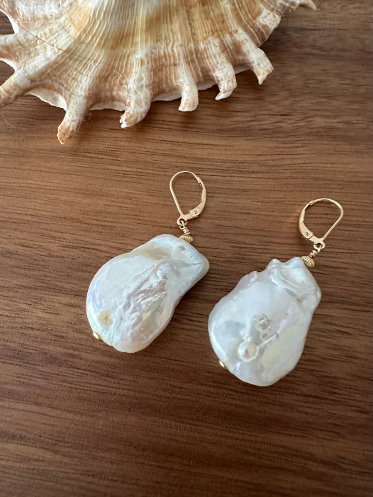 a twinned pair of white large earrings with gold ear hooks on a wooden background. there is a shell in the upper edge of the frame