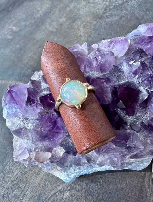 a round opal ring in gold iwth four beads at the four directins is on a piece of brown leather sitting on a piece of amethyst  on grey wood