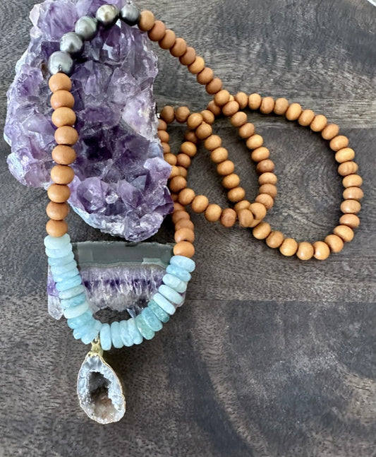 a necklace with blue aquamarine, sandalwood and tahtian pearl beads with a raw geode is on a purple crystal on a wooden background