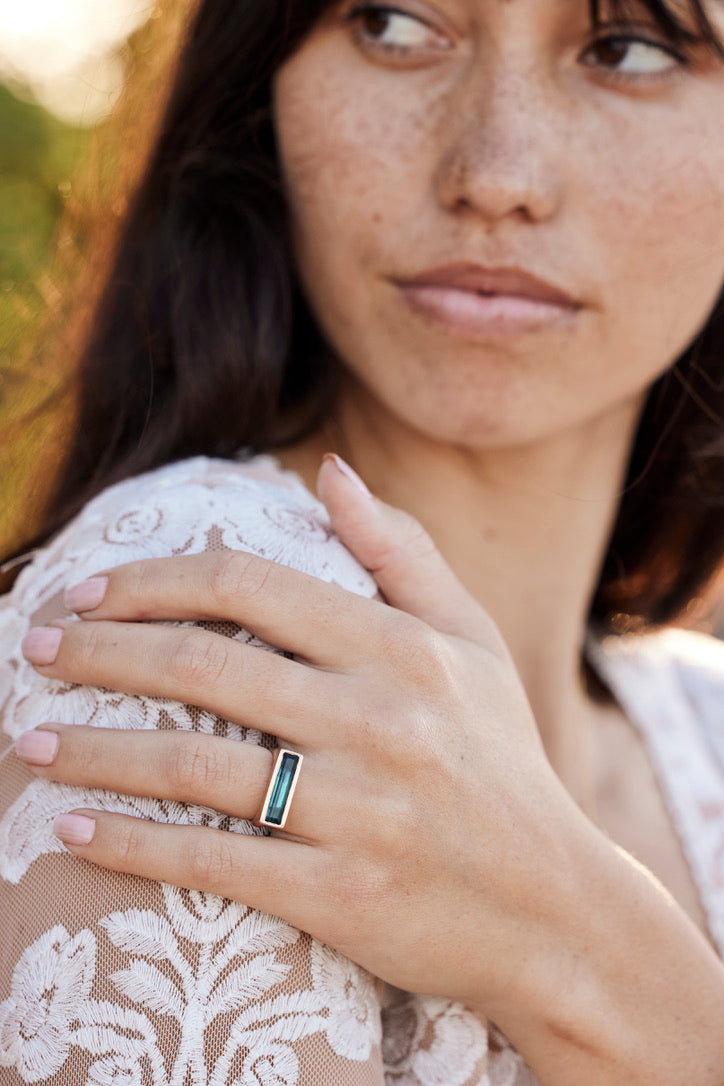 a pretty woman with brown hair is out of focus but her hand which is on her shoulder is showing a blue rectangular ring that is a horizontal orientation