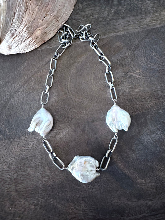 a large necklace with three large white cloud shaped pearls set between sterling silver links lays on a wooden background with a shell in the upper left corner
