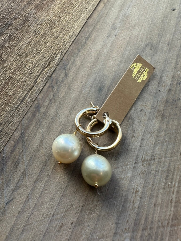 large whitish pearls with a sublte golden tone on little huggie hoops on a brown craft tag with a golden logo that says tide and tied on a wooden background