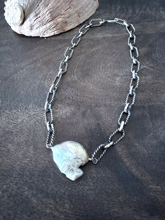 a large necklace with silver links and a large cloud shaped pearl in the center lies on a wooden background with an abalone shell in the upper left corner