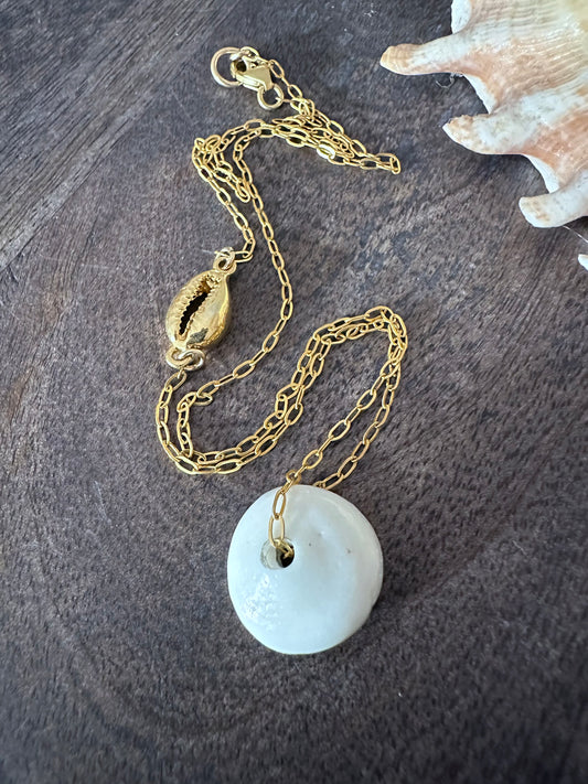 a necklace with a large white puka shell and a gold cowrie shell on a wooden background with a shell in the upper rt corner.