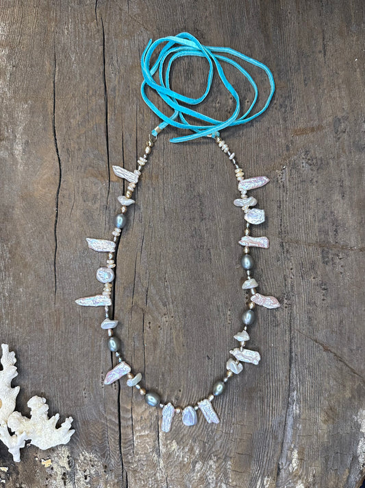 a necklace with turquoise suede tails and grey and silver pearls sits on a wooden background with some coral in the lower left par tof the frame