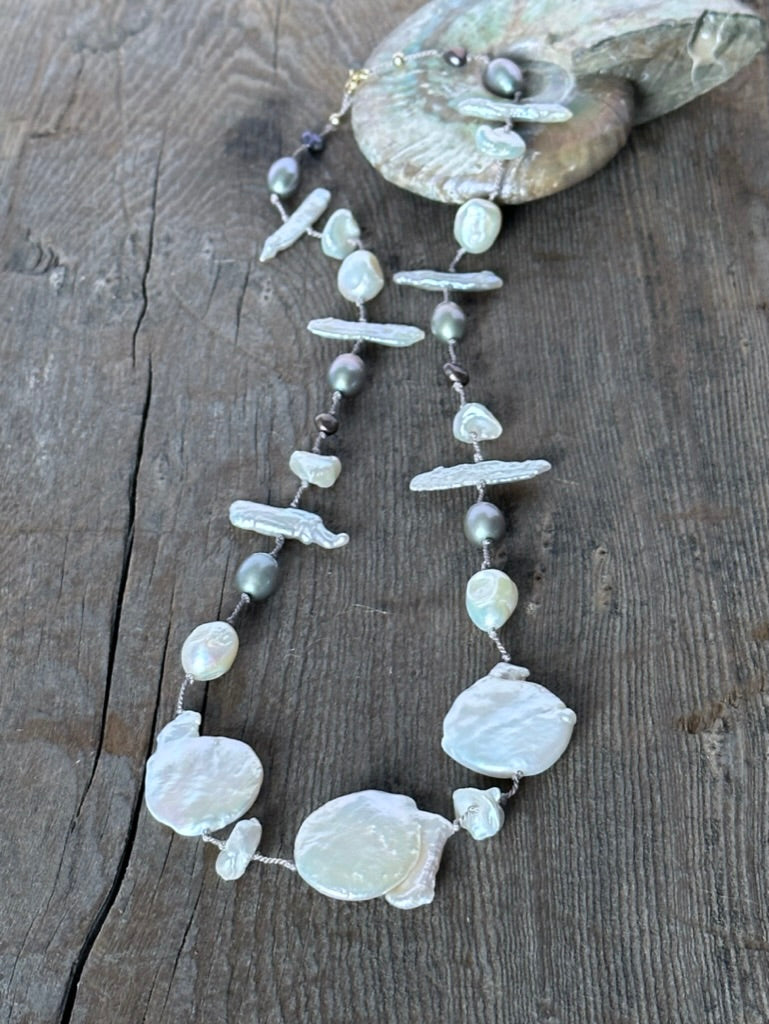 a necklace with three large cloud shaped white pearls and smaller white and grey perals on a wooden background wth a fossilized nautalus shell blurred out inthe background