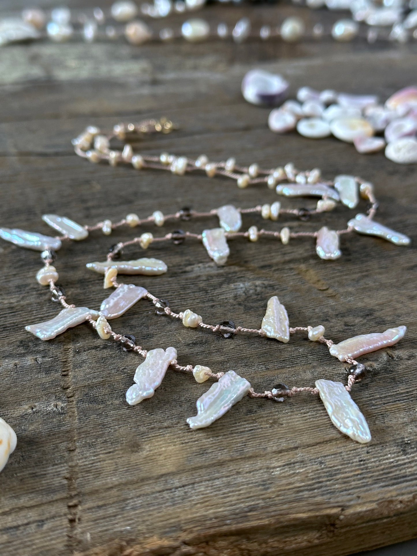 a pinkish silk necklace on a wooden background with odd shaped pearls and brown faceted beads. there are some puka shells in the upper right corner and lower left edg of the frame