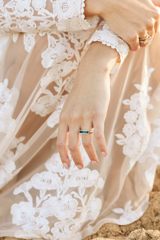 a woman hand in a lace dress with a ring on it. the ring is a long rectangular blue and rosegold ring with an east west presentation