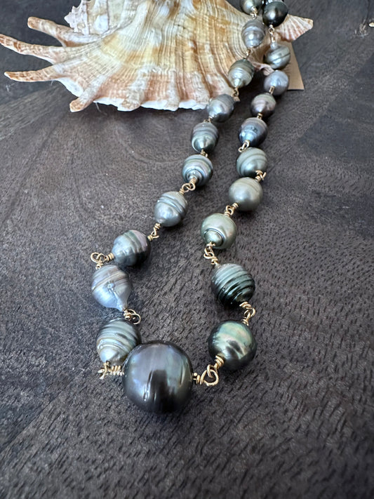 a string of black pearls that have been wire wrapped together to make a neckalce lays on a shell on a wooden backdrop
