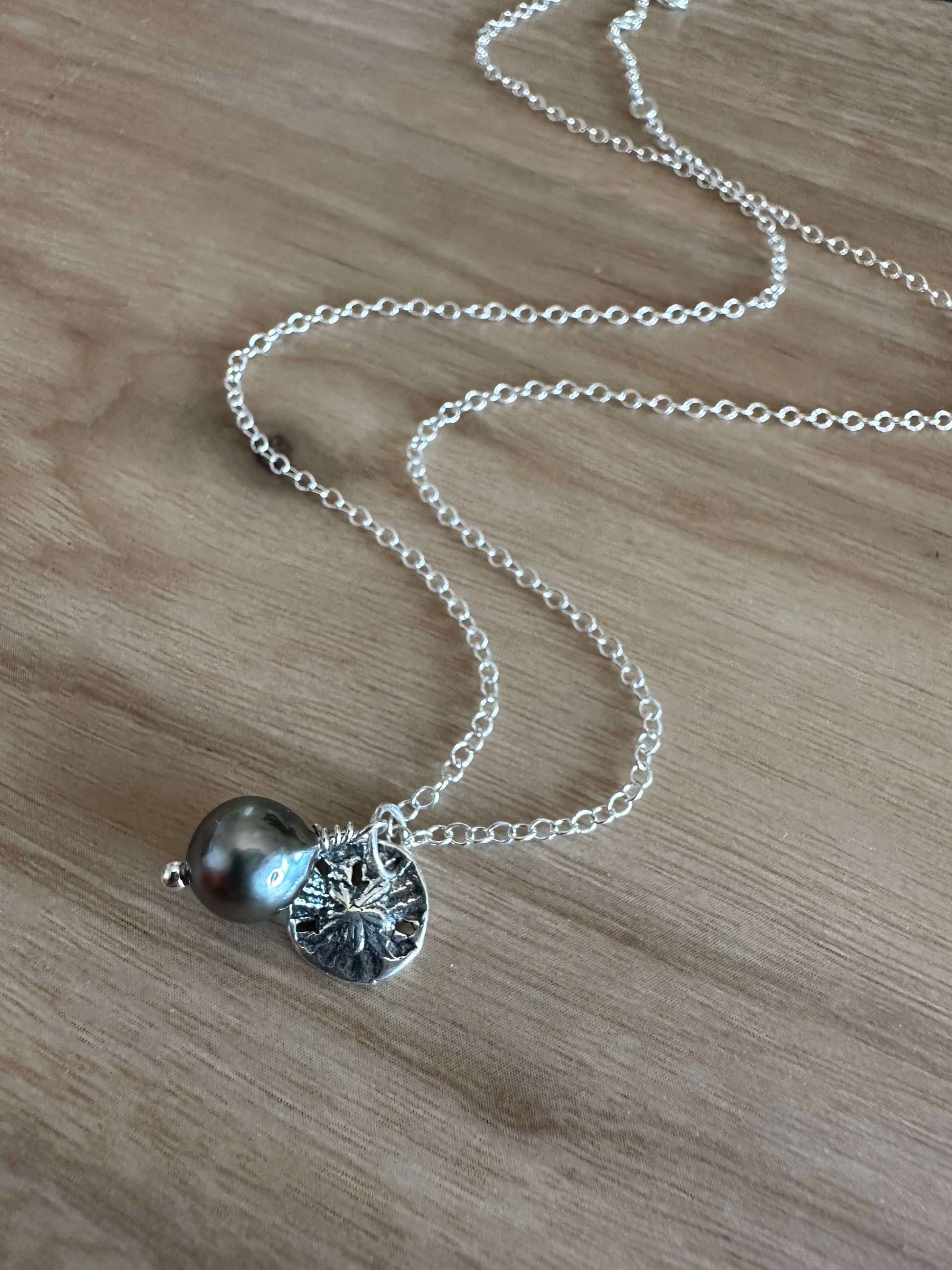balck tahitian pearl ans silver sandolar charm pendant on a silver chain lays on a wooden background