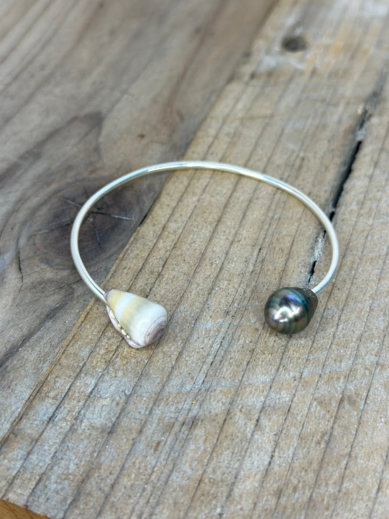 a silver cuff bracelet with a yellow banded shell on one side and a black pearl on the other side sitting on a wooden background