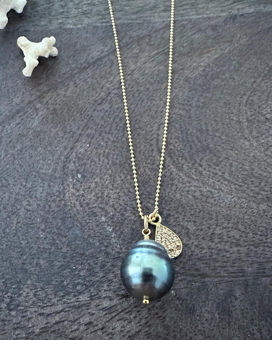 a light blueish black tahitiaon pearl with a diamond pave teardop pendant on a gold chain on a wooden background with small white coral in the upper left corner.