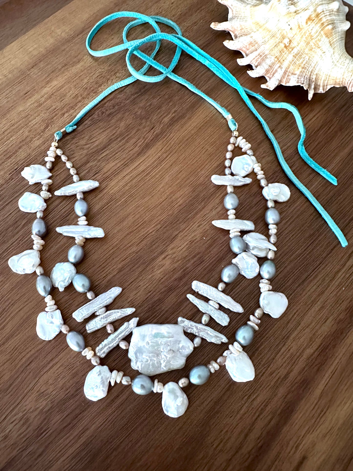 a necklace with two strands of white and grey long and round pearls and a large cloud shaped pearl in the center with a turquoise tail on a wooden background. a seashell is in the upper right corner