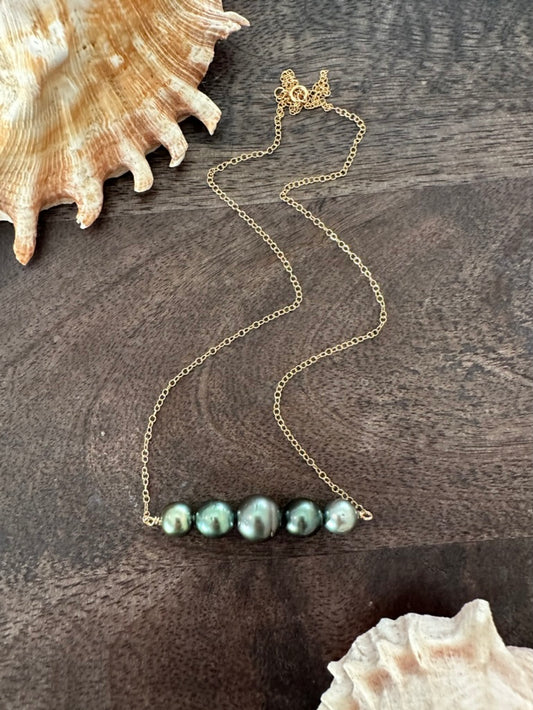 a necklace with five black pearls in a row on a gold chain on a wooden background wtih shells in the upper left and lower right corner.
