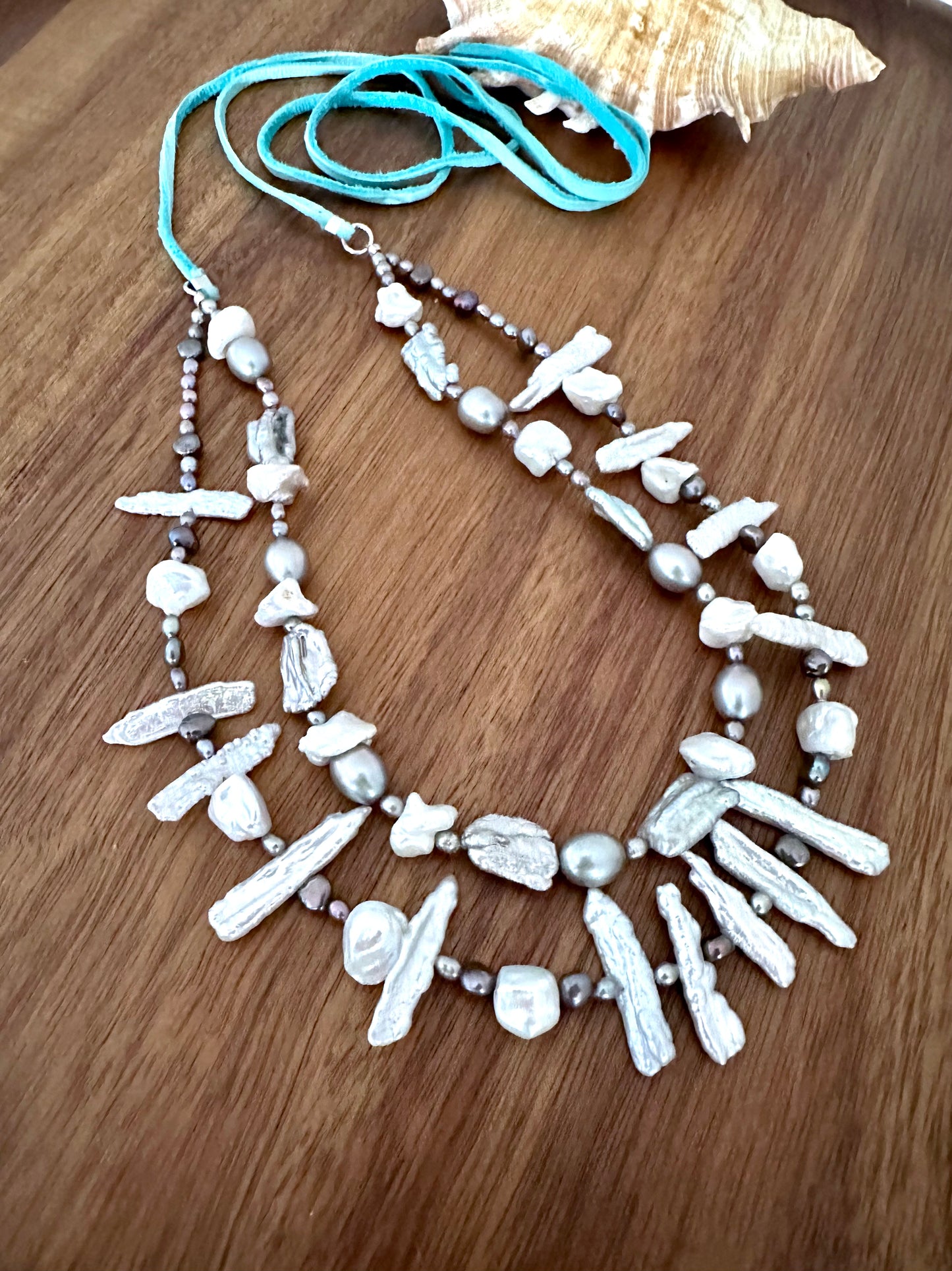 a necklace with two strands of white and grey long and round pearls with a turquoise tail on a wooden background. a seashell is in the upper right corner