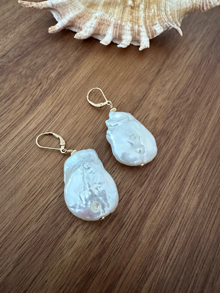 a twinned pair of white large earrings with gold ear hooks on a wooden background. there is a shell in the upper edge of the frame