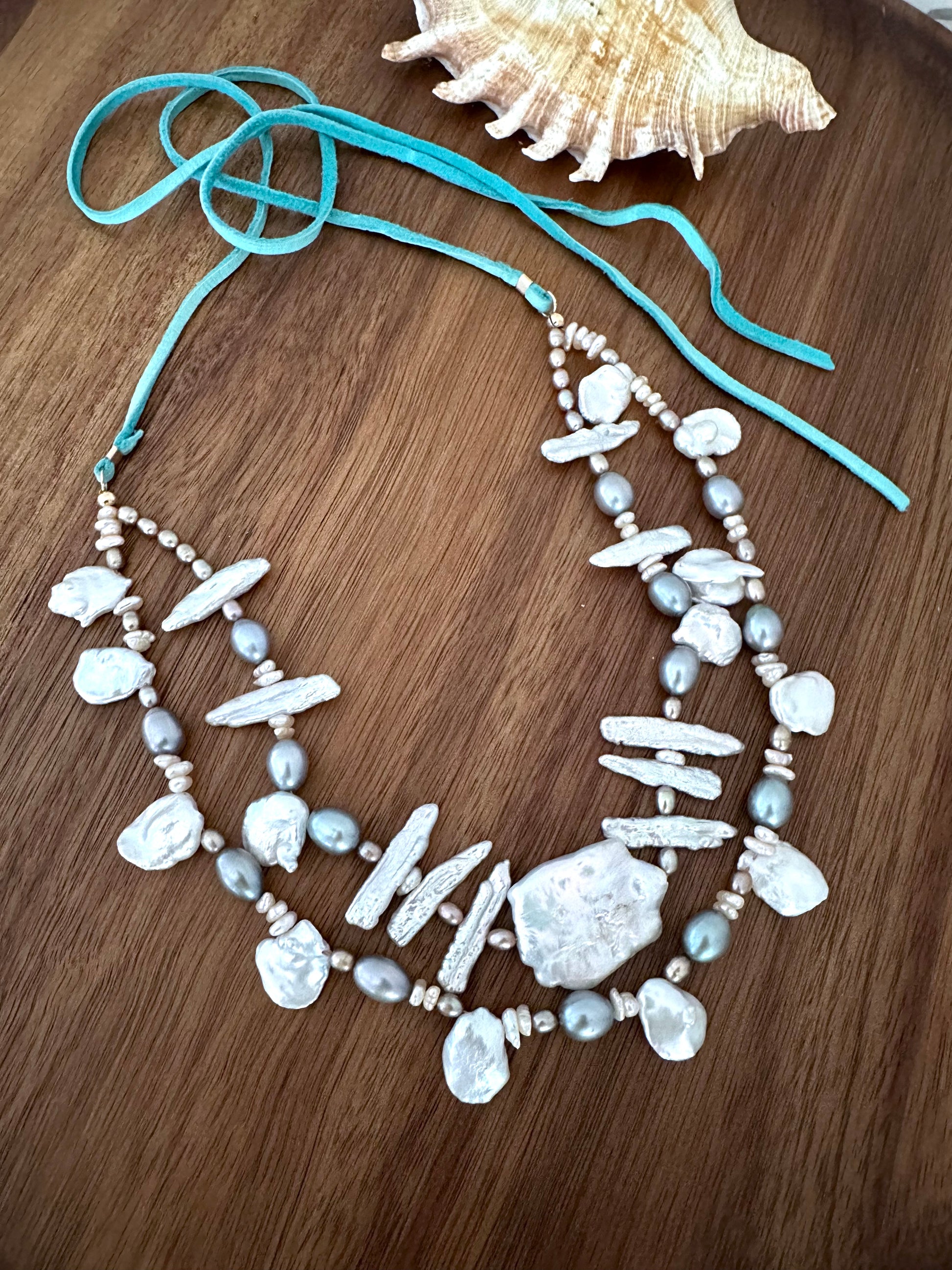 a necklace with two strands of white and grey long and round pearls and a large cloud shaped pearl in the center with a turquoise tail on a wooden background. a seashell is in the upper right corner