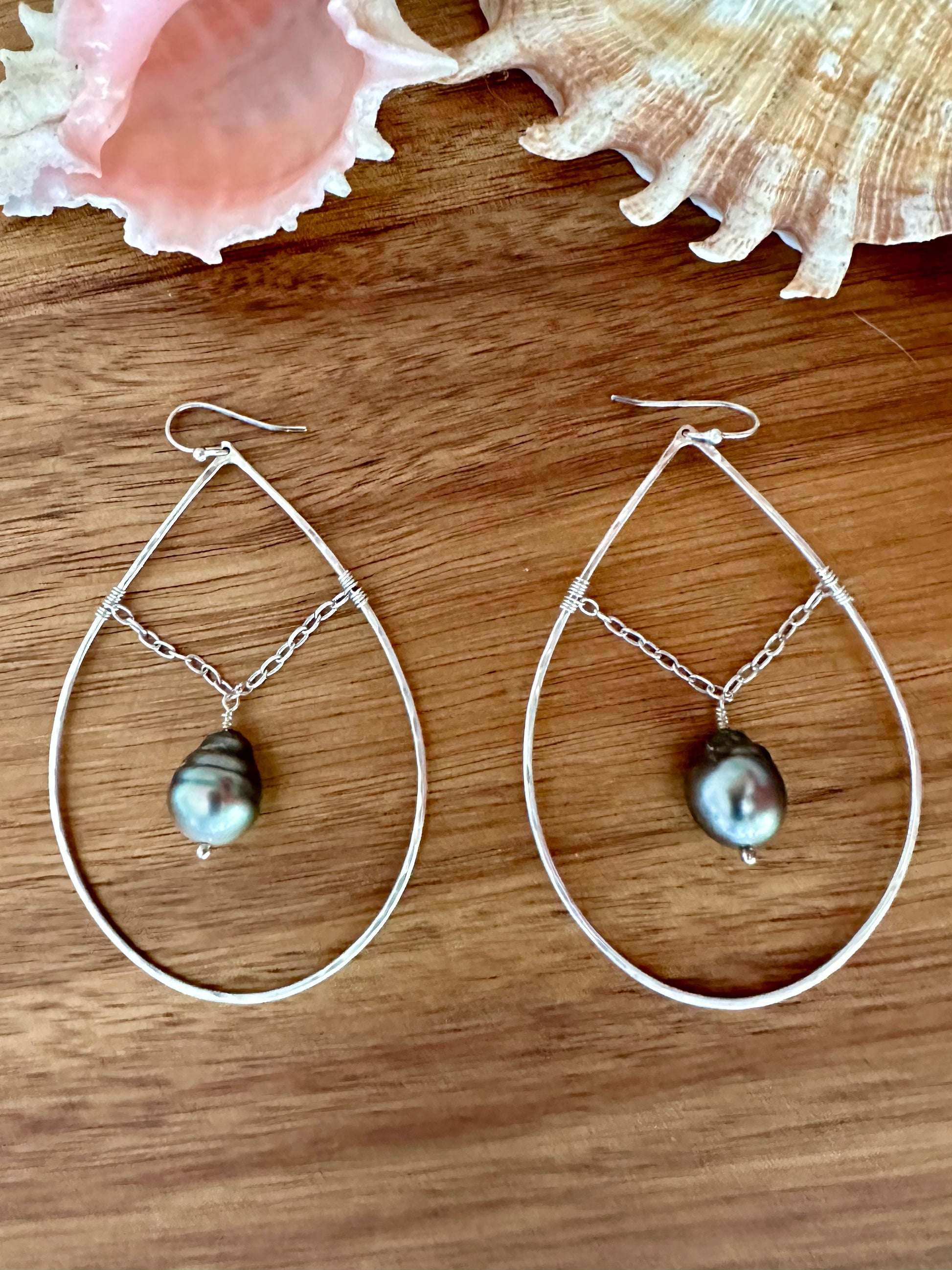 a pair of sterling silver earrings with a chain suspending a black pearl. thye are on a brown wooden background with two shells in the upper part of the frame