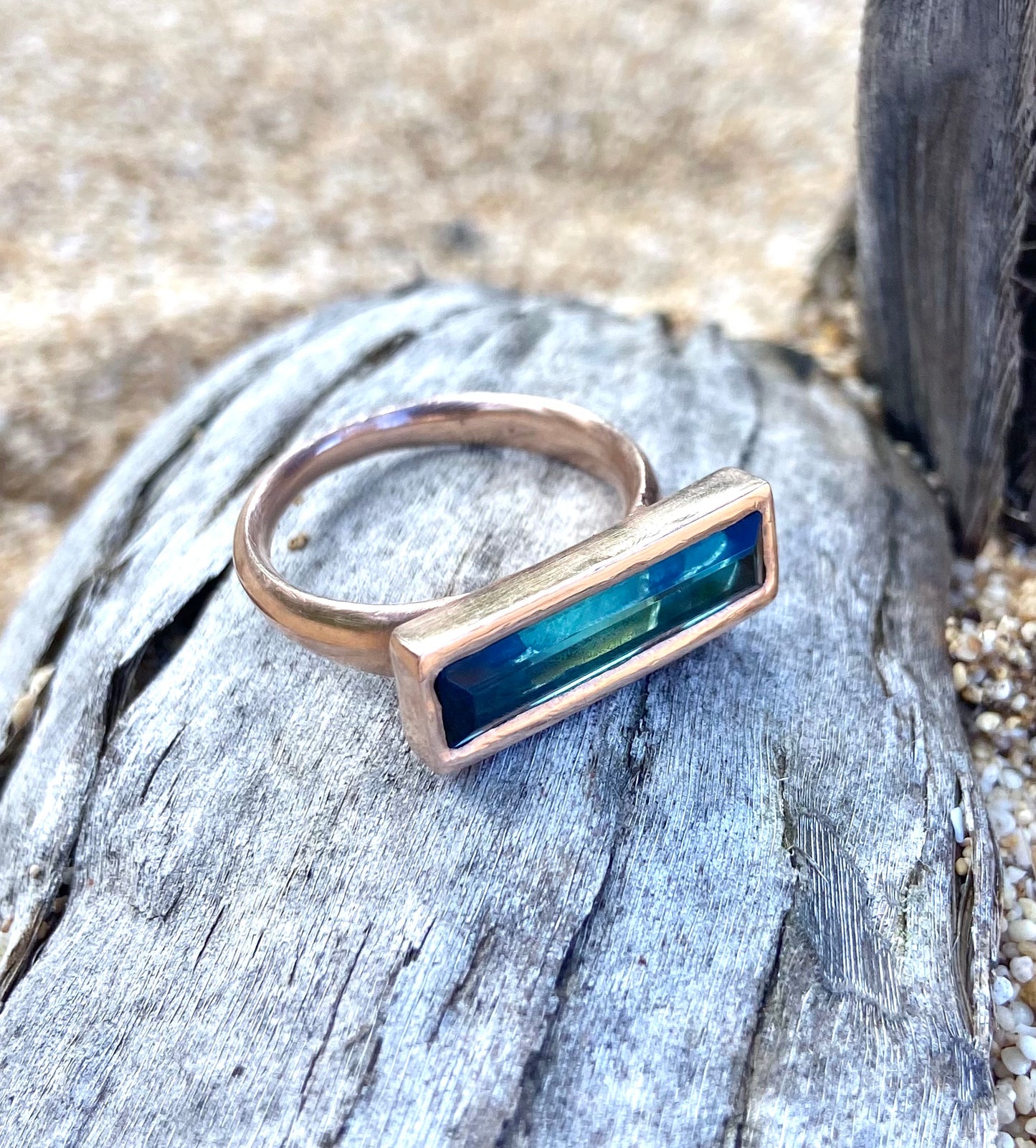 a blue rectangular stone in a rose gold setting sits on a piece of drift woond in the sand