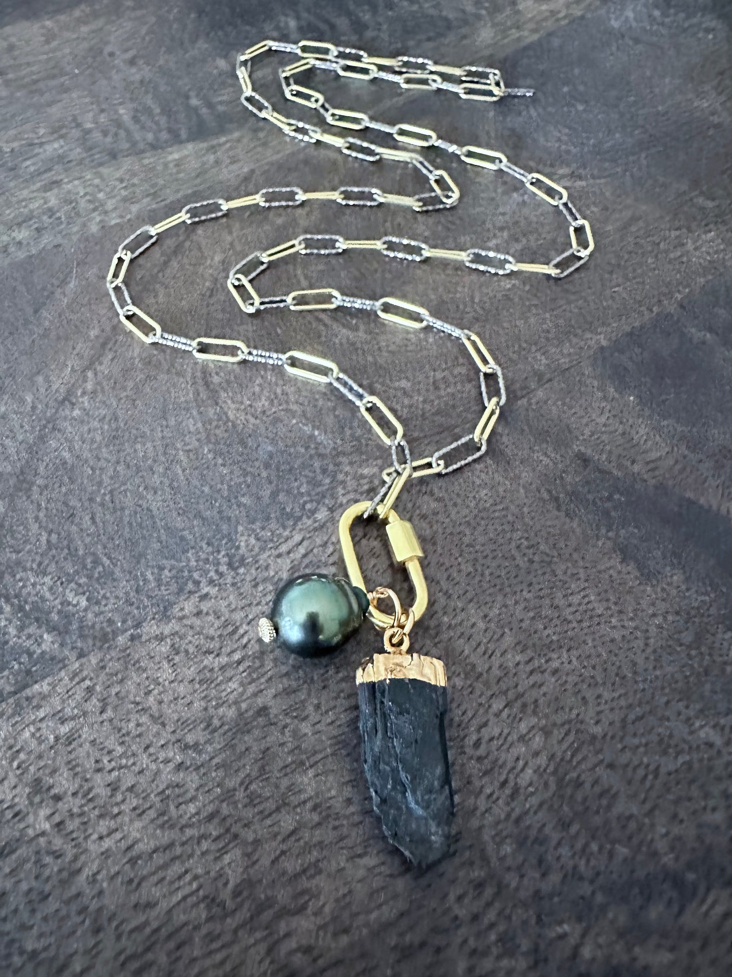 a long necklace wth mixed metal chain with a gold carabeaner and a large black pearl and a raw black tourmaline pendant on a wooden background