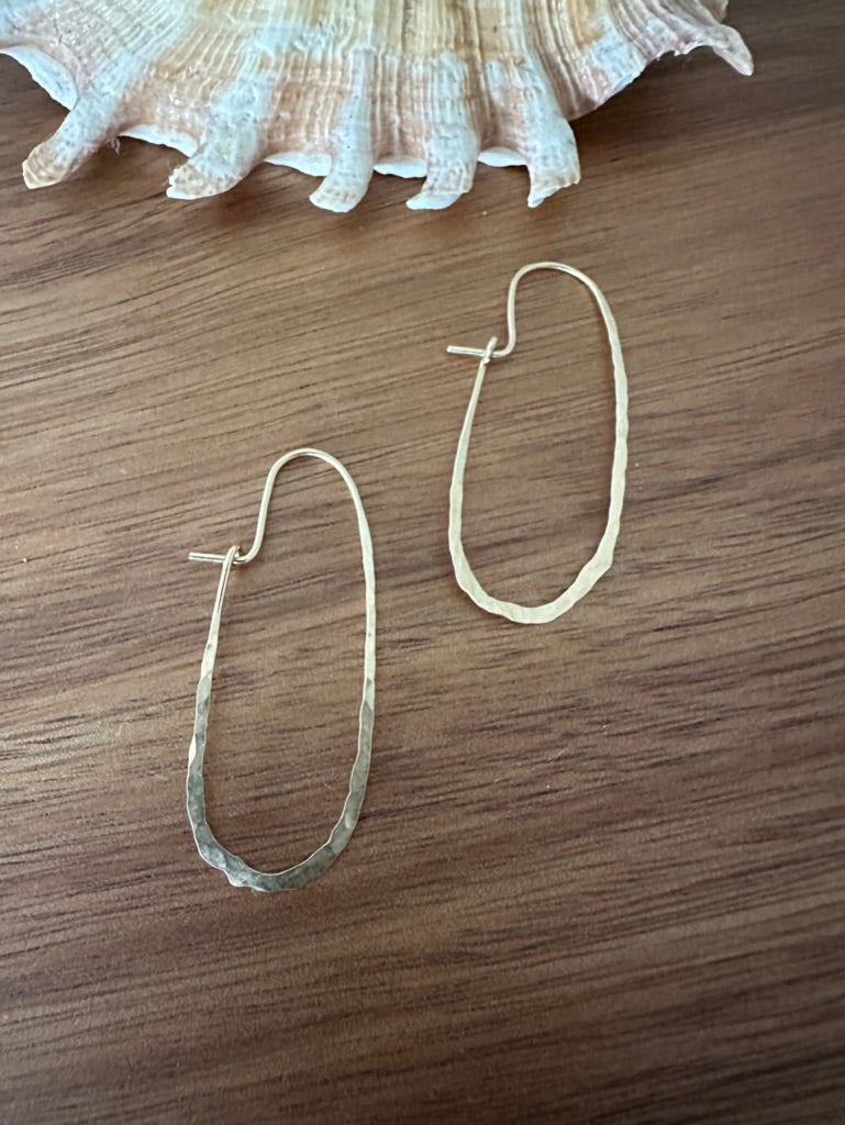 elongated gold hoops made out of a single piece of wire. it sits on a brwon wooden backdrop with a shell in the upper edge of the frame