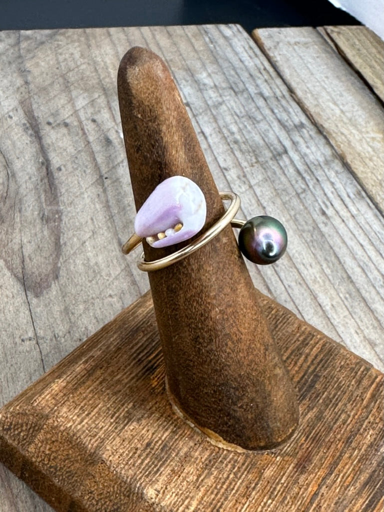 a ring on a wooden ring stand with a purple shell and a tahitian pearl on a wire that twists around the ring holder. you can see some sand in the opening of the shell