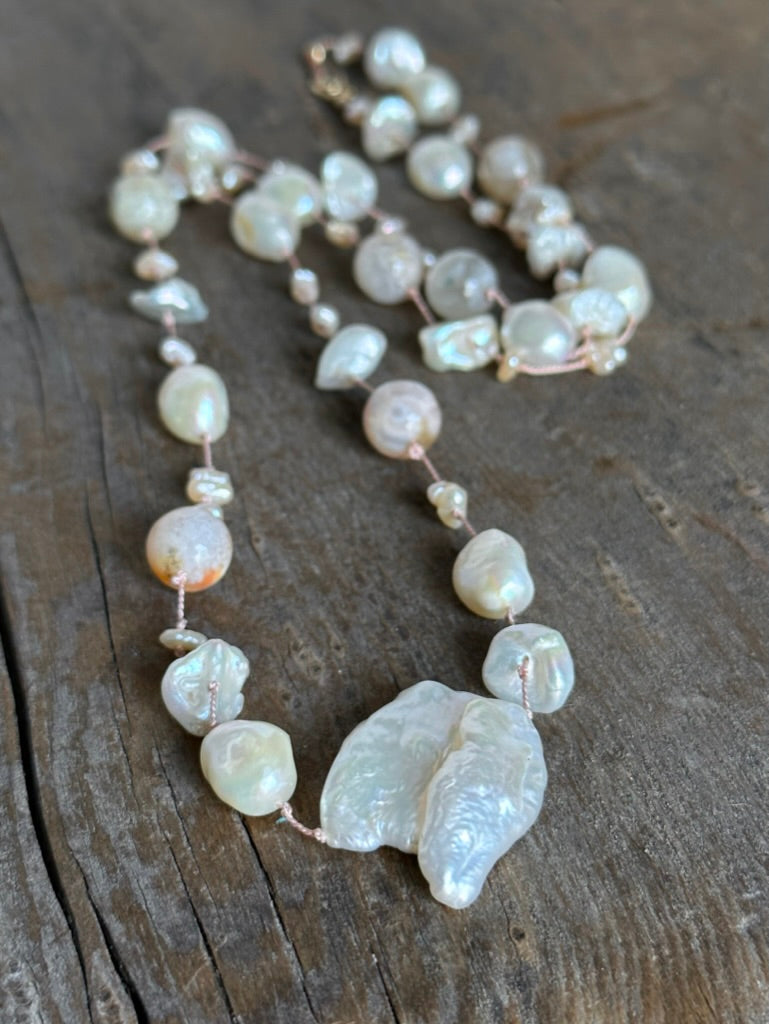 a pinkish silk necklace on a wooden background with a large cloud shaped pearl and round pinky flower agate beads