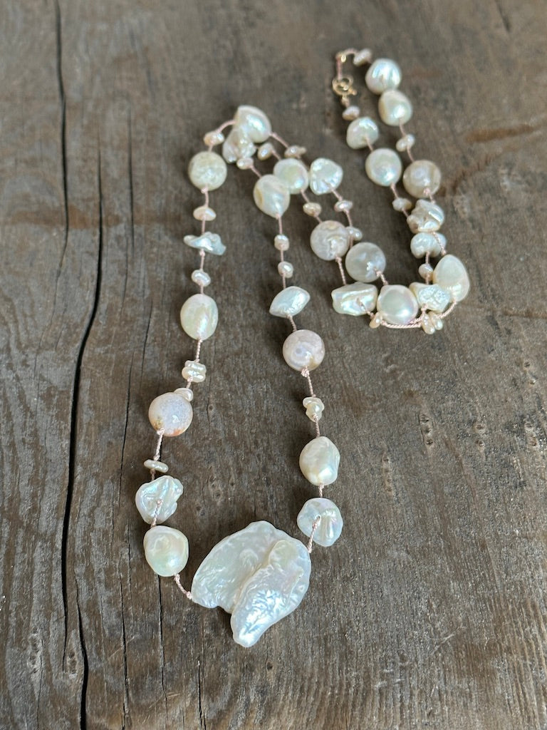 a pinkish silk necklace on a wooden background with a large cloud shaped pearl and round pinky flower agate beads  