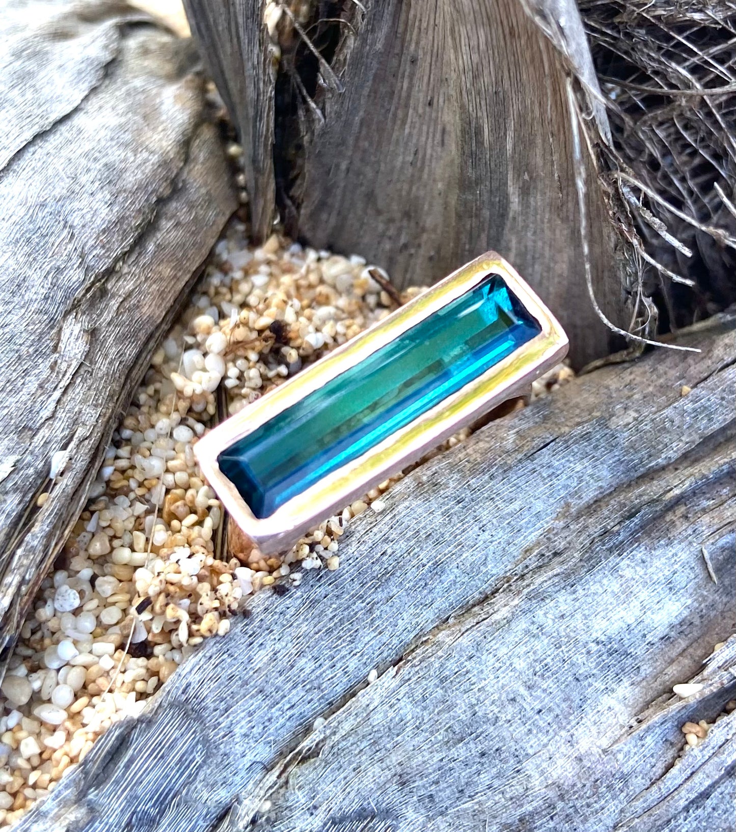 a rectangular ring sits in sand. the stone is very blue.