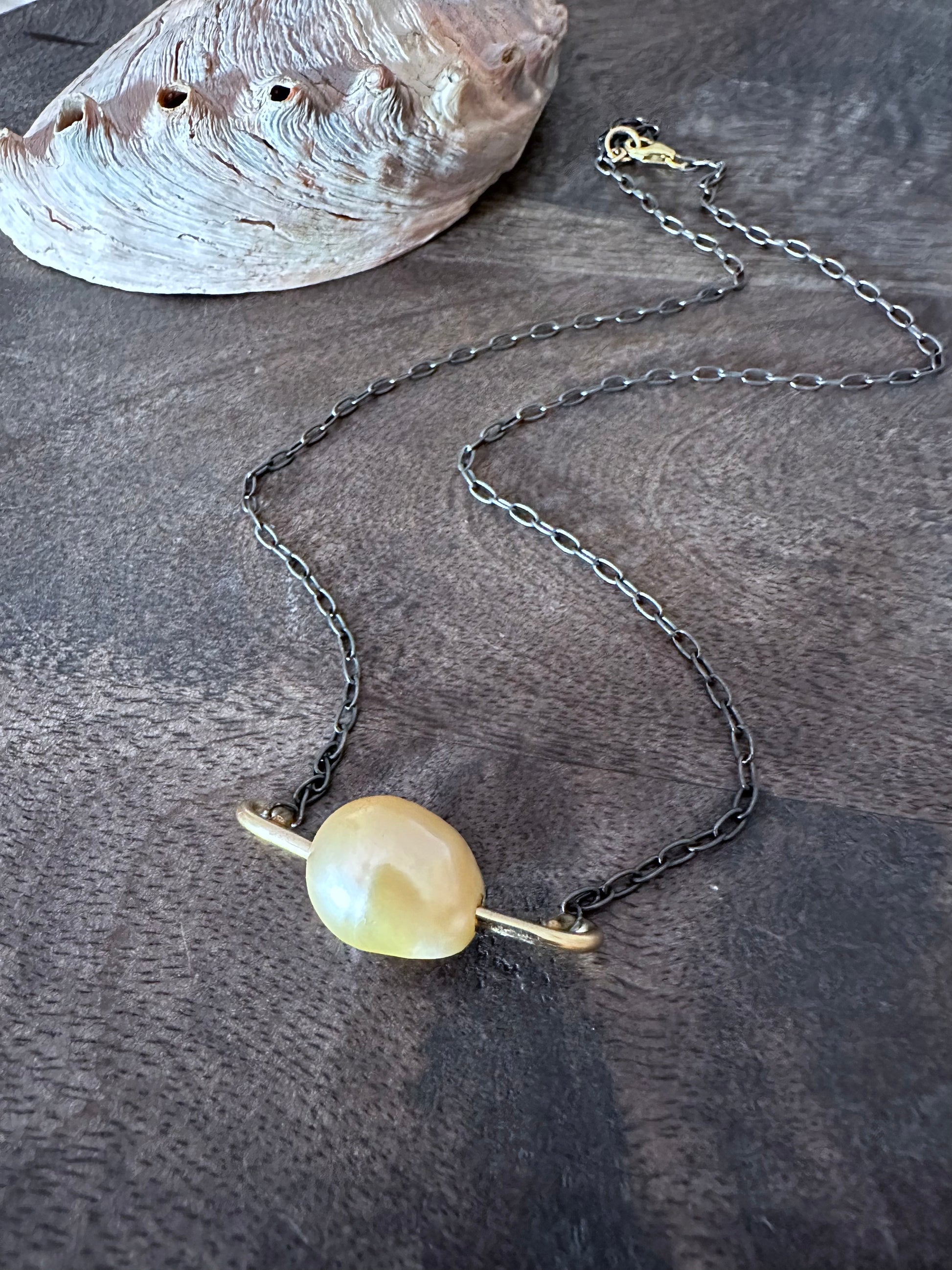 a necklace on a dark wooden background. the necklace has a yellow/pink pearl with a gold bar gong through it on a darkened silver chain. there is an abalone shell in the upper left corner.