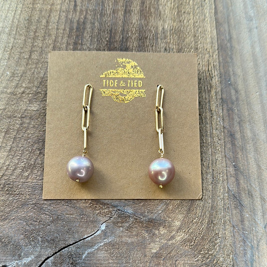 A pair of pink earrings on a brown card with a gold logo on it. it is laying on a brown wooden background