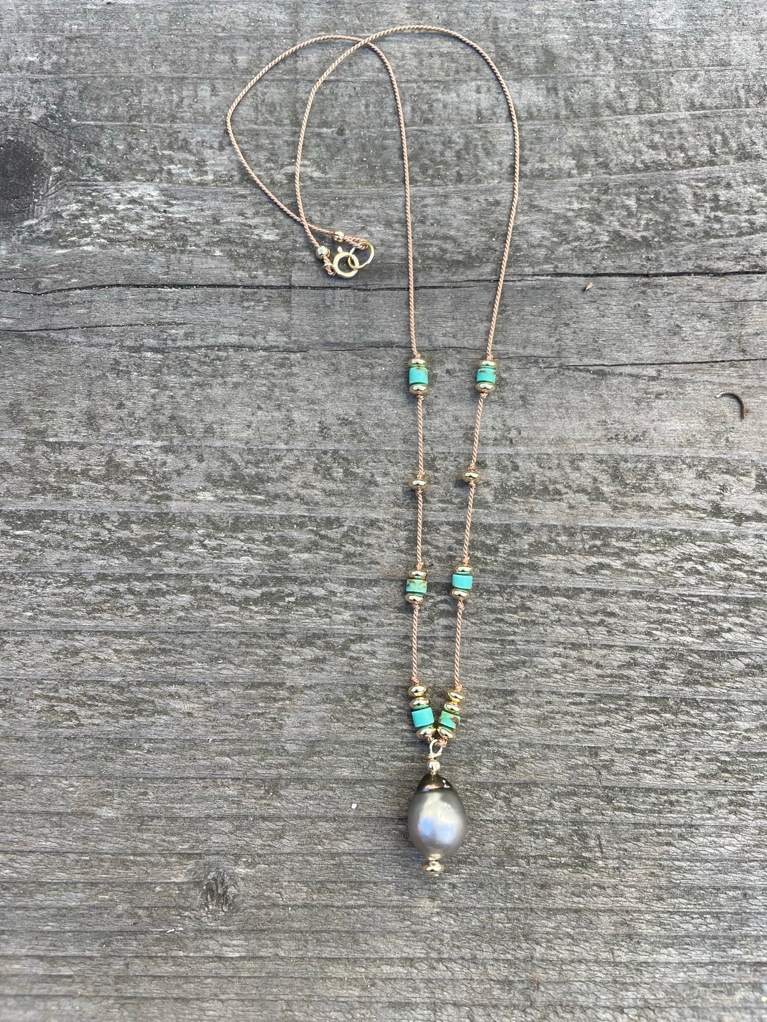 a necklace with turquoise and gold beads and a center pendant of a tahtian black pearl sit on a wooden background