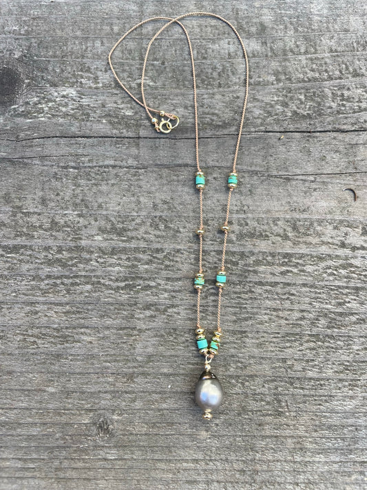a necklace with turquoise and gold beads and a center pendant of a tahtian black pearl sit on a wooden background