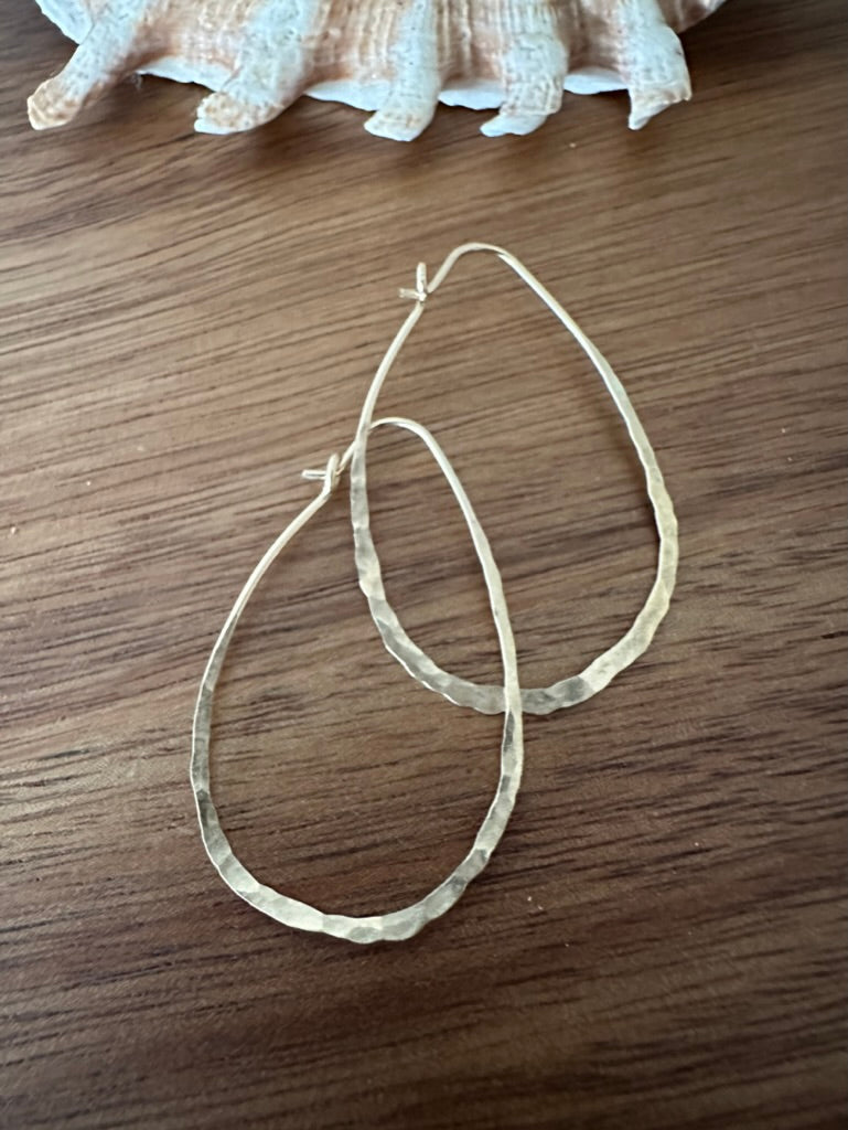 a pair of gold hoop earrings that are hammered and made from a single continuous wire. they are shaped like a lare teardrop or kind of like and avocado. they sit on a wooden backdrop with a shell in the upper part of the frame