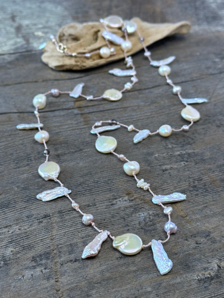 a pinkish silk necklace on a wooden background with odd shaped pearls and round pinky golden pearls with a piece of drift wood blurred out in the background