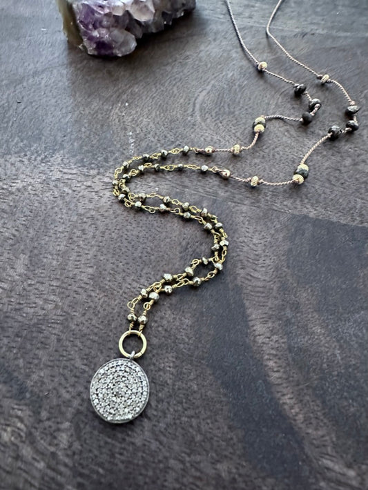 a necklace with a round pendant of diamond pave in sterling silver hangs off of a gold and silvery pyrite chain and gold beads on silk ona wooden background iwth a crystal in the upper left corner.