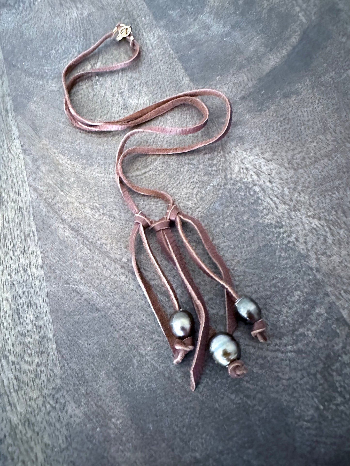 a soft suede necklace with fringes and three tahitian pearls ont he fringes on a light grey wooden background.