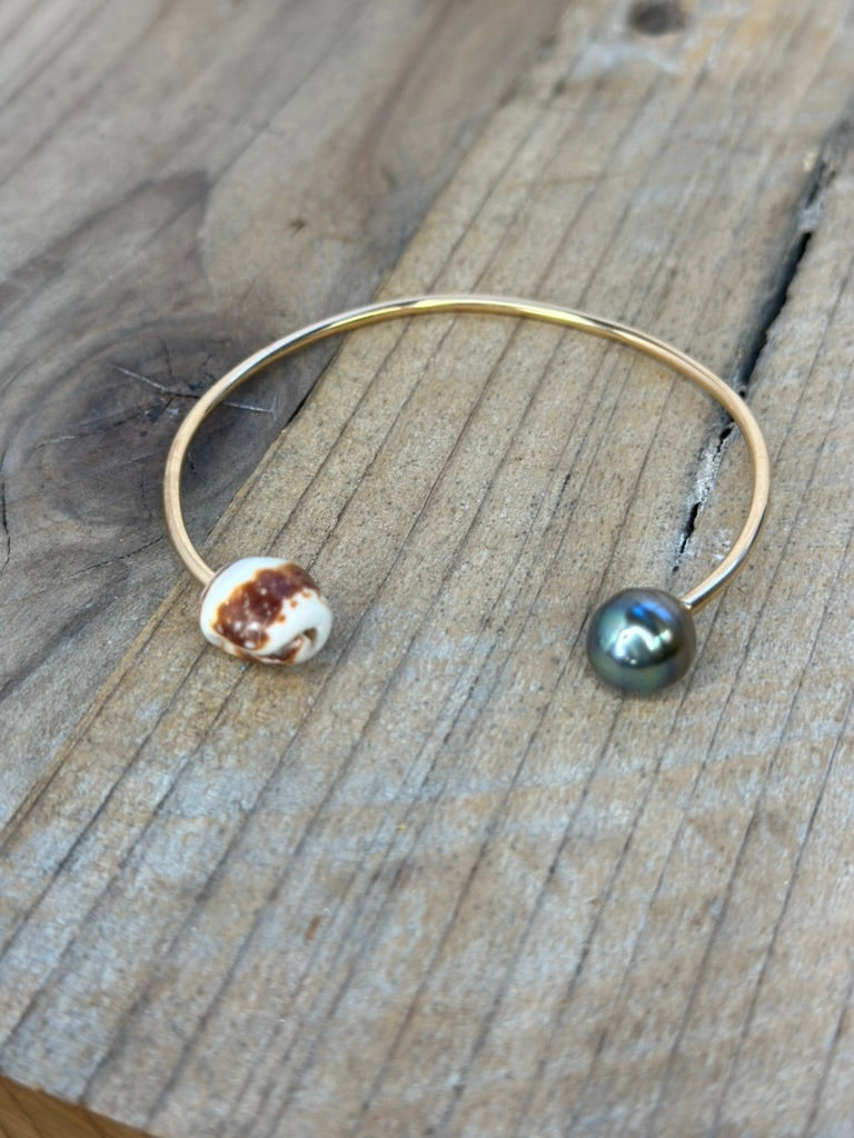 a cuff bracelet in gold with a brown and white shell on one side and a balck pearl on the other