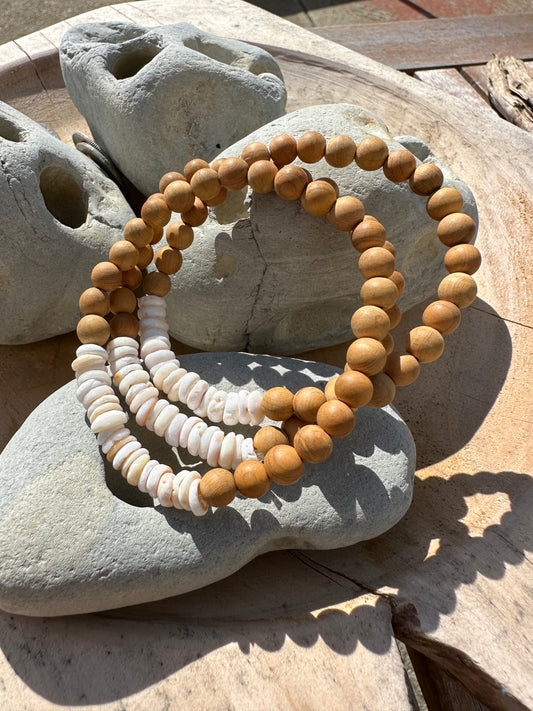 three sandalwood beaded bracelet with puka shell on grey rocks sitting in a wooden bowl in the sun.