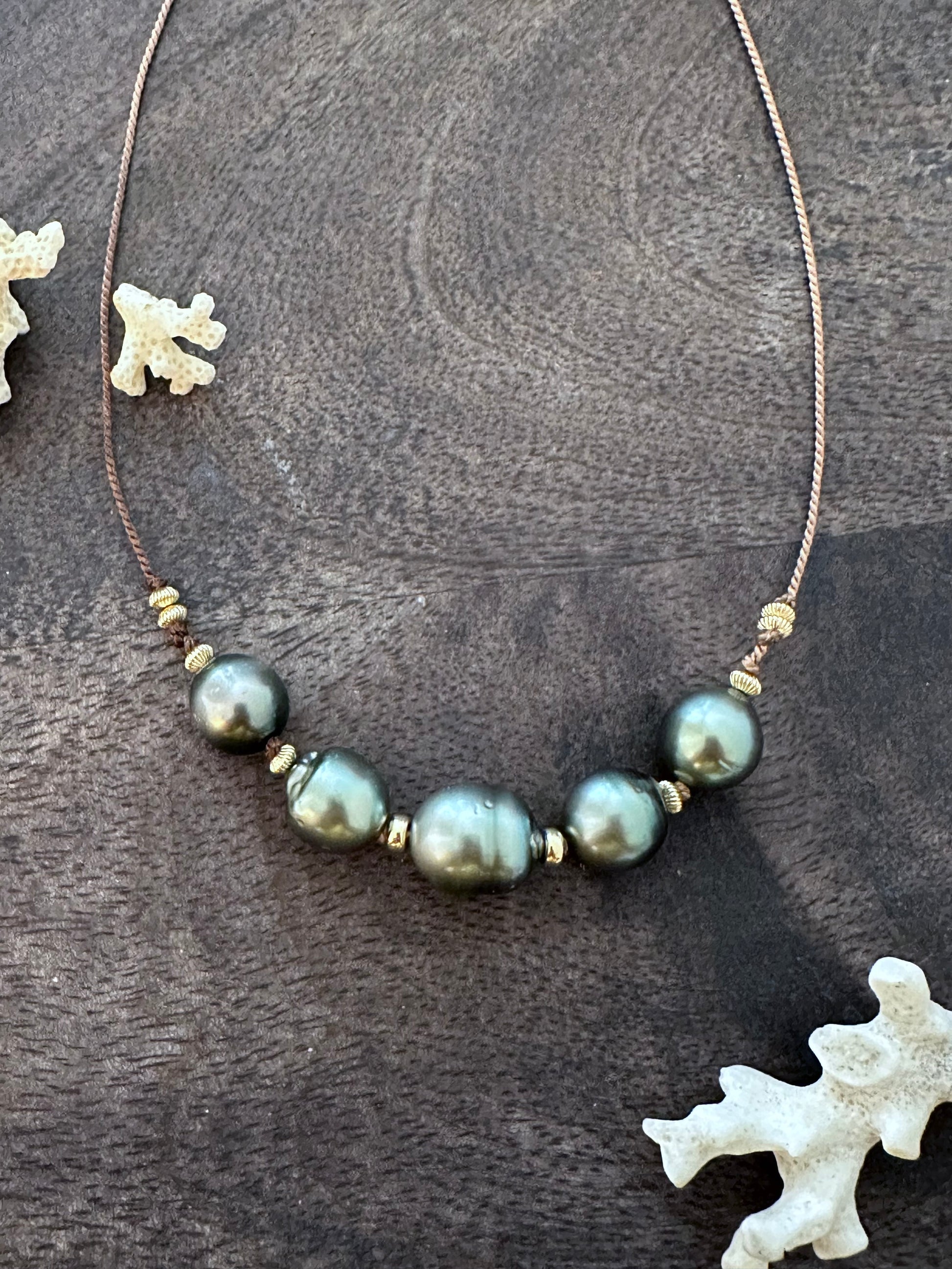a necklace with 5 large tahitain pearls on a tan silk string with gold filled beads on a wooden background with white coral in the lower right corner and upper left corner.
