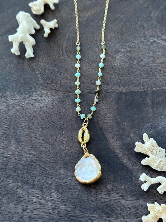 a necklace with a gold cowrie and a white flat pearl with gold edges on a turquoise rosary chain is on a grey wooden backdrop. there are white coral pieces in the upper left andlower right corner