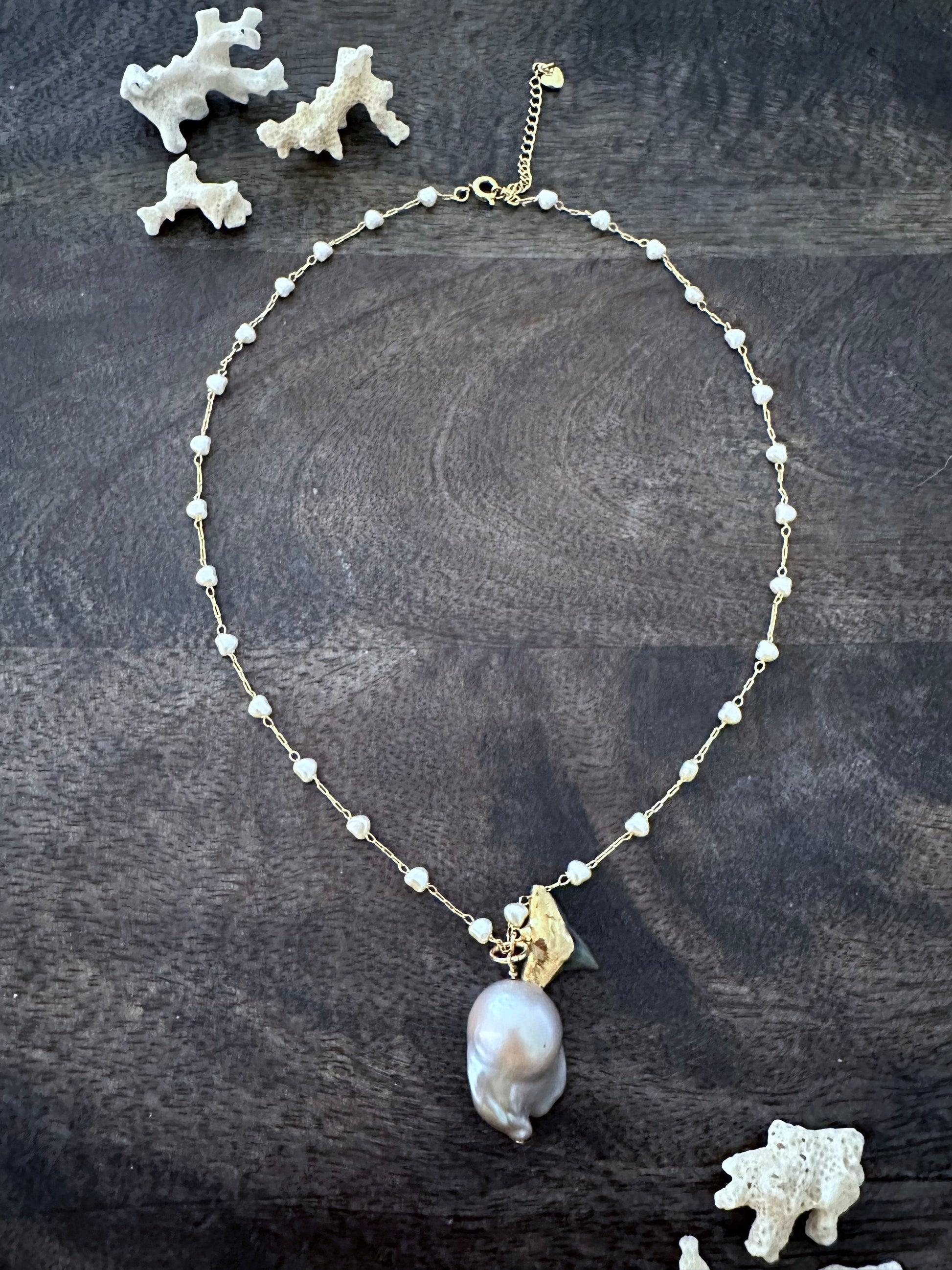 a white pearl necklace with a large grey oddly shaped pearl and a gold vermeil black sharks tooth on a grey wooden background. there are white coral pieces in the upper left andlower right corner