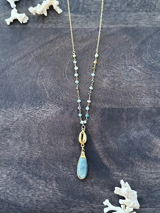 long blue pendant with a gold cowrie shell on a blue rosary chain on a wooden backgroudn. there are white coral branches in the upper left and lower right corners
