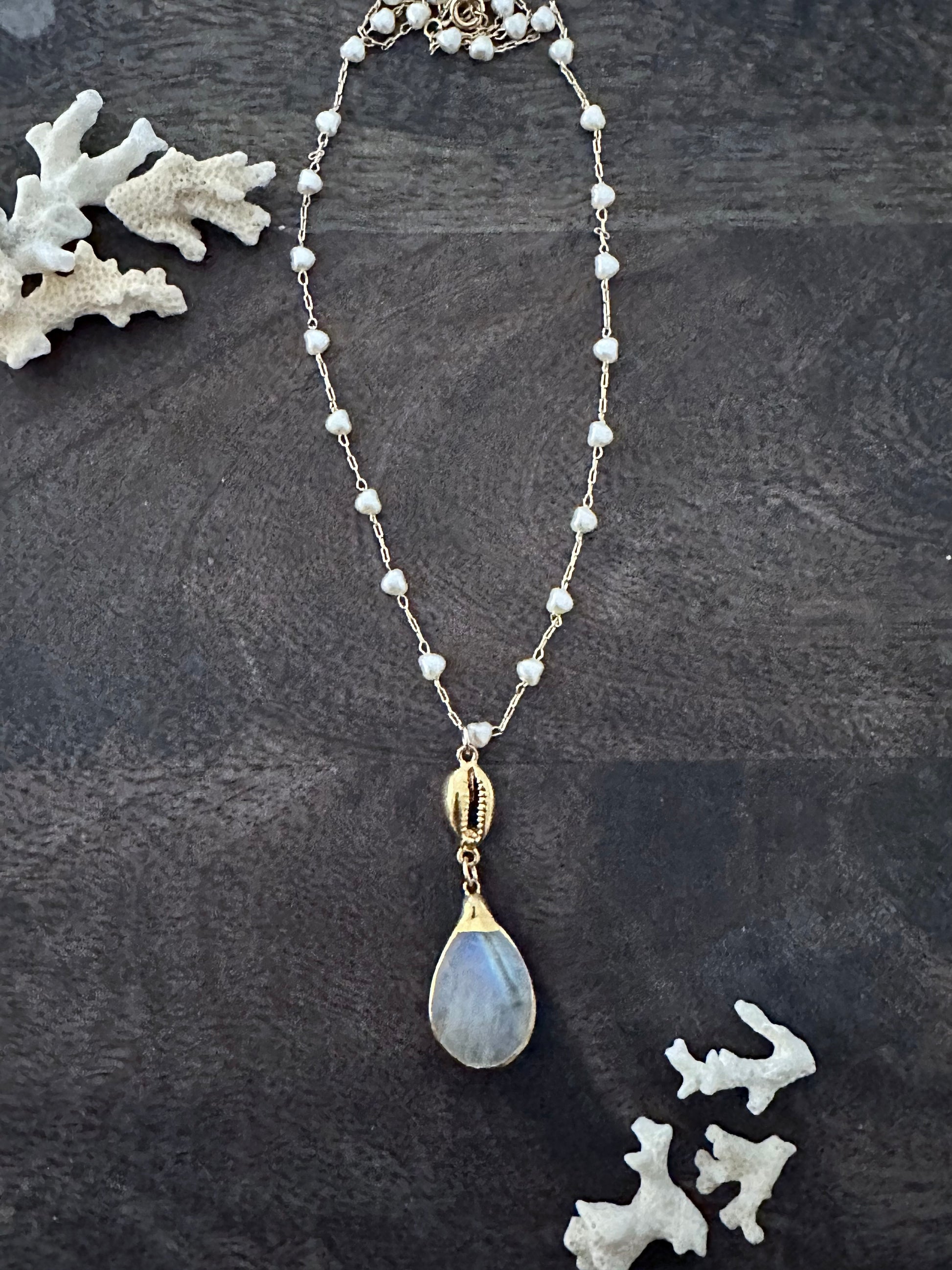 Necklace with a rainbow moonstone pendant with a gold cowie shell above it on a 18" necklace with white heart shaped pearls on a wooden background with white coral pieces in the upper left and lower right corners