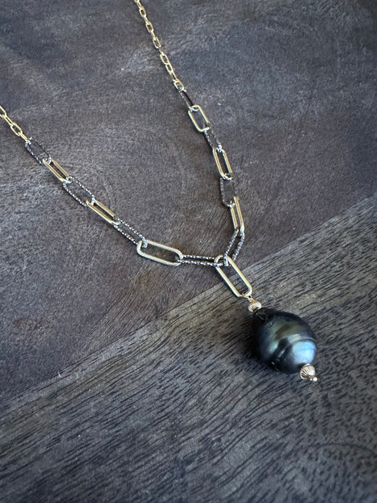 A large black tahitian pearl on a mixed silver and gold chain on a grey wooden background