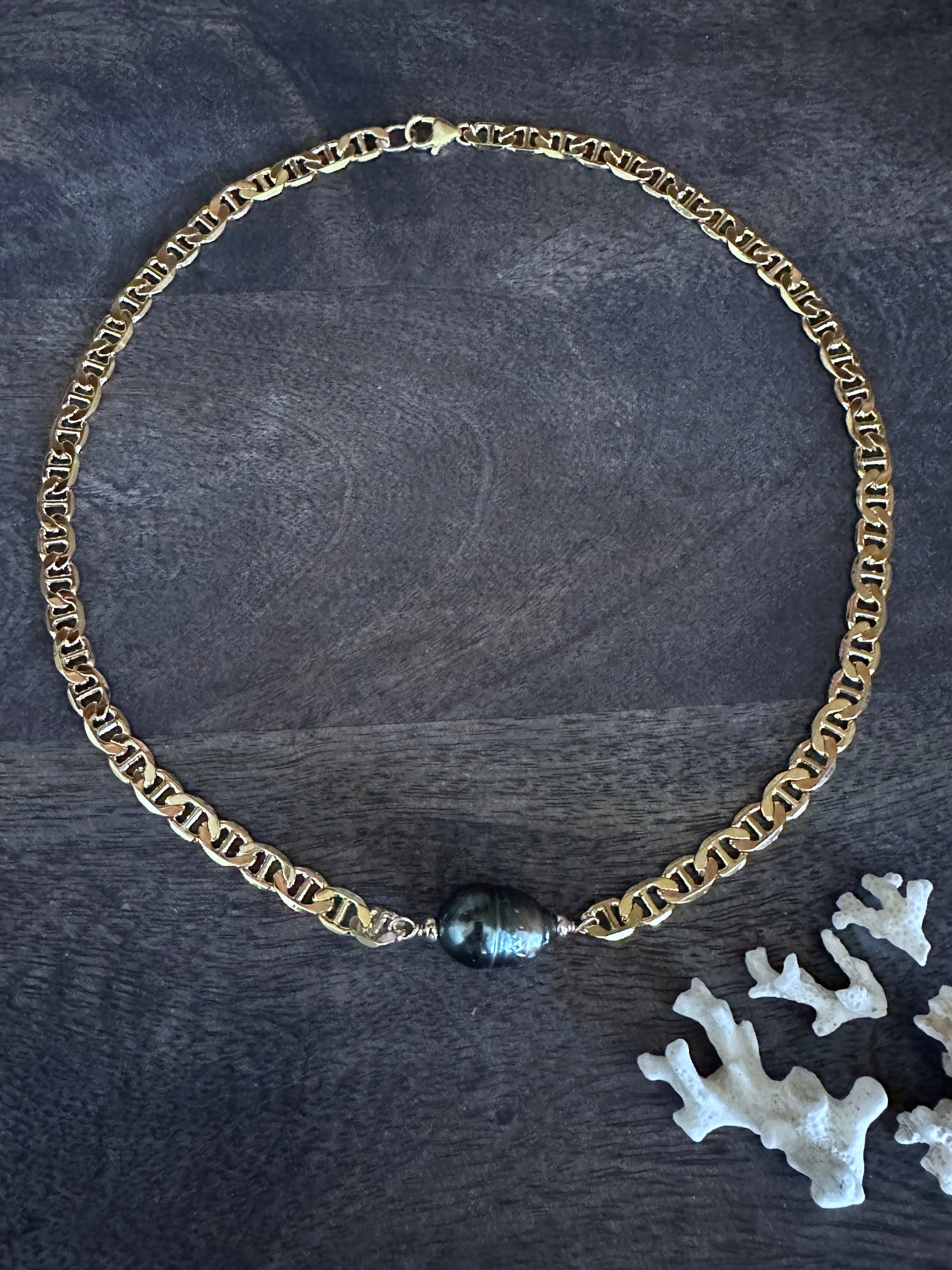 a large flat gold neckalce lays in a circle on a grey wooden background with a large tahitian pearl as the center piece. there are small pieces of white coral in the lower right hand corner of the image.