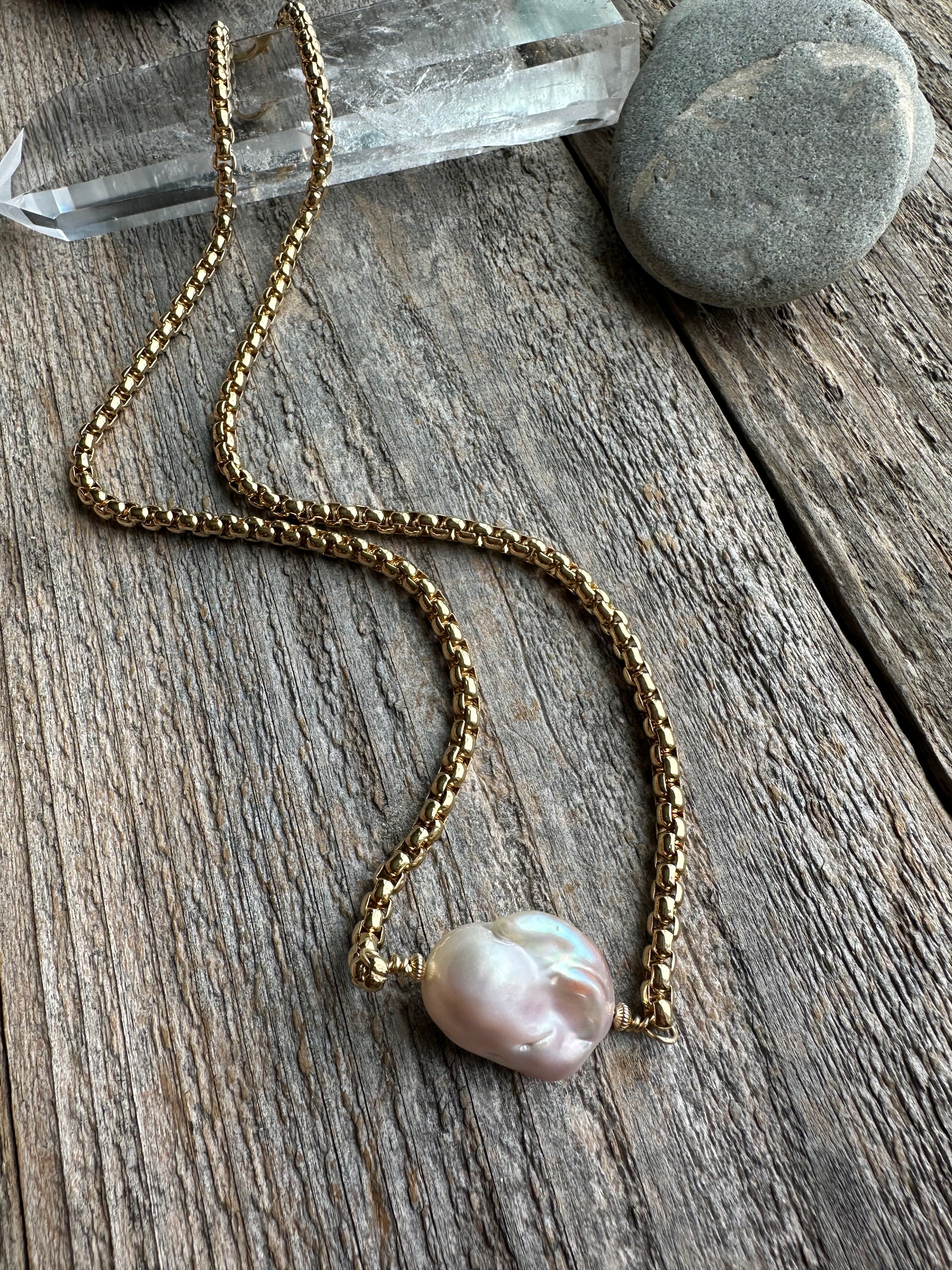On wooden background is a gold necklace with an unusually shaped pearl laying on top of a quartz crystal and rock in the upper part of the image.
