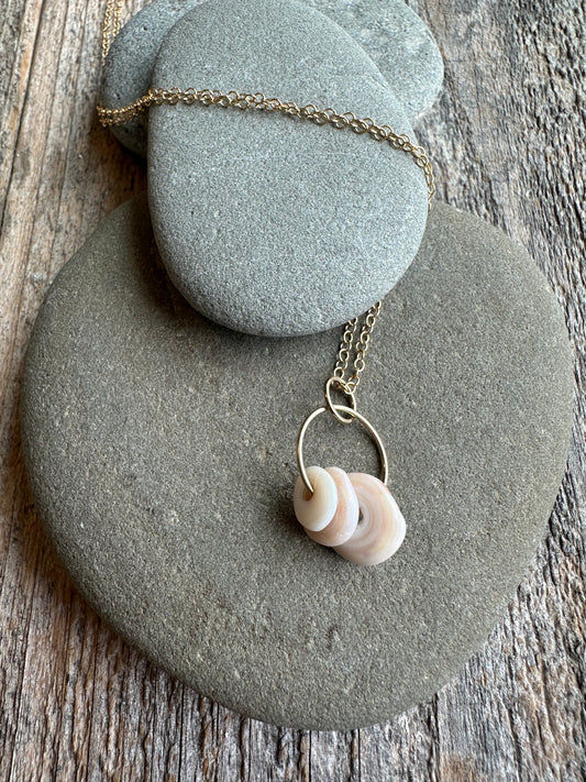 On wooden background there is three rocks with one stacked on top of the other two. Layed on top and wrapped in one of the rocks is a gold chain necklace with 3 golden puka shells.