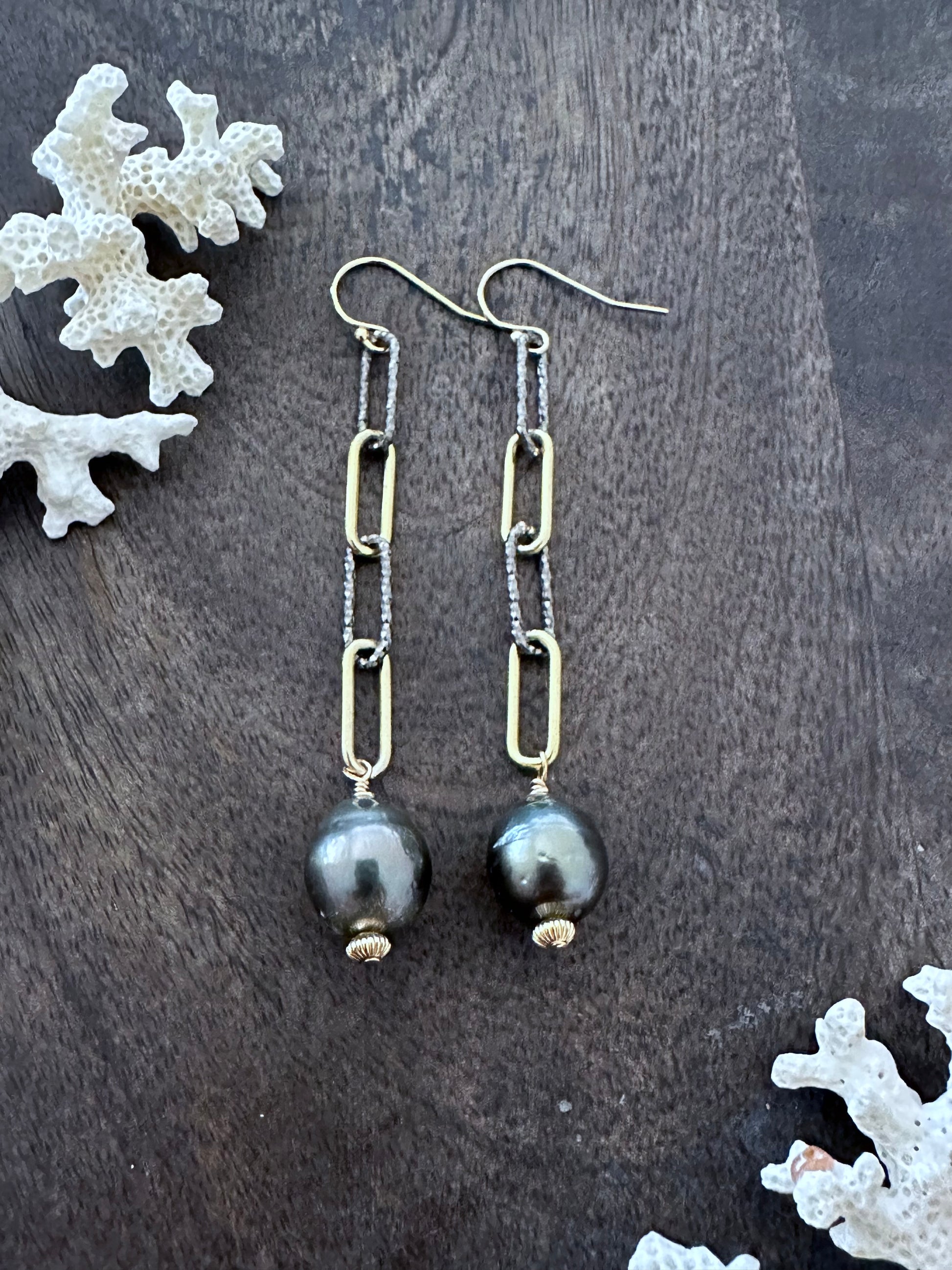 a pair of earrings on a grey wooden back ground. they are alternating links of silver and gold with a singel tahtian black pearl at the bottom. there are white coral pieces inthe upper left and lower right corners of the image