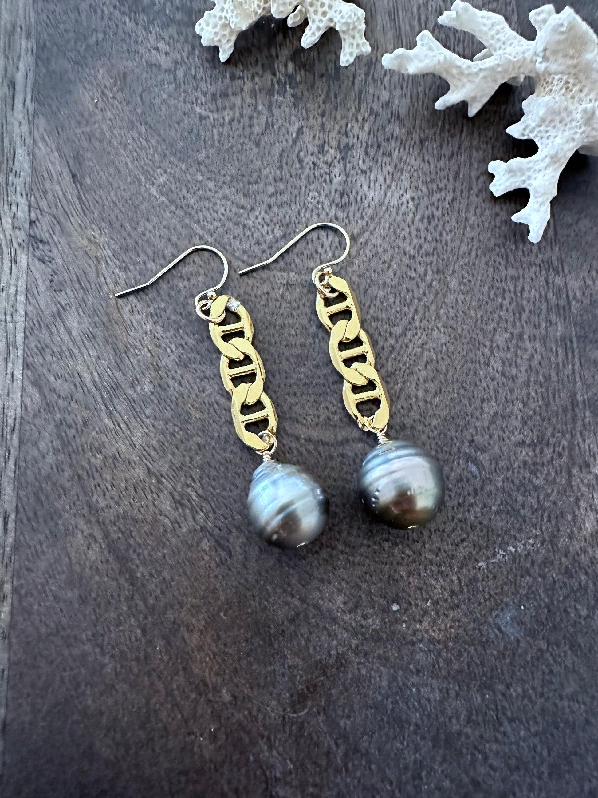 a pair of earrings on a grey wooden background with large flat curb chain and tahitan pearls on the ends. there3 are pieces of white coral in the upper left corner